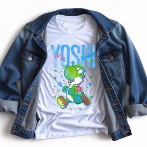 May include: A white t-shirt with a graphic of Yoshi, a green dinosaur character, and the word "YOSHI" in blue. The shirt is styled with a blue denim jacket. The design includes colourful paint splatters.