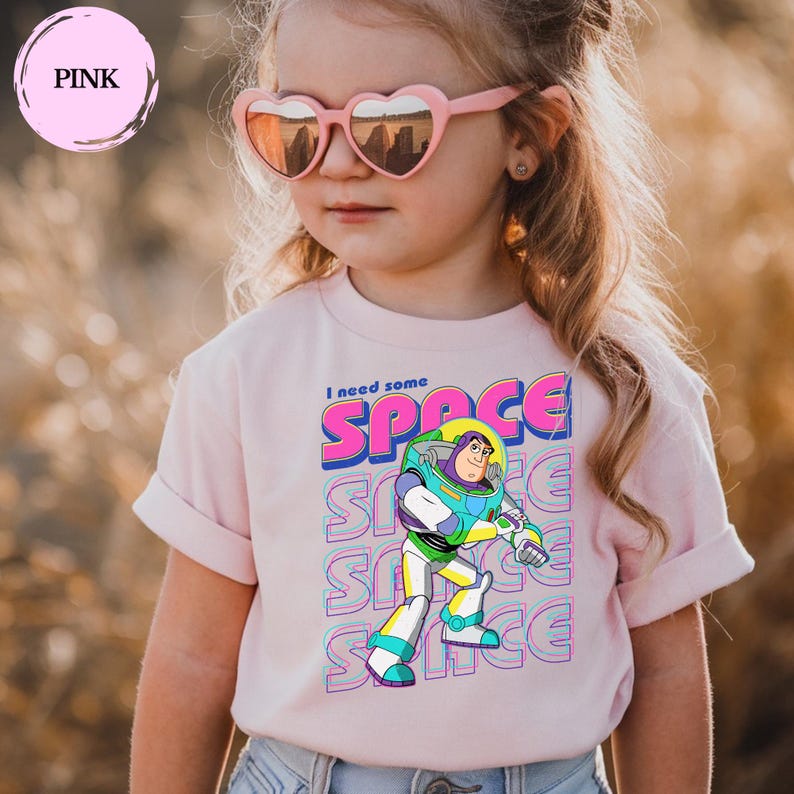 May include: A light pink t-shirt featuring Buzz Lightyear from Toy Story with the text "I need some SPACE". The design includes the word "SPACE" repeated vertically. The shirt is worn by a child.