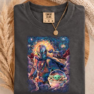 May include: Dark grey Comfort Colors t-shirt with a vibrant illustration of The Mandalorian and Grogu set against a starry night backdrop. The tee has a crew neck and short sleeves. A gold necklace with a circular pendant is draped on the shirt.