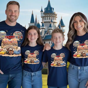 May include: Four people wearing navy blue t-shirts with a Cars-themed design. The design features a road, cartoon cars, and the word "Cars" in a Mickey Mouse head shape. They are smiling in front of a castle.