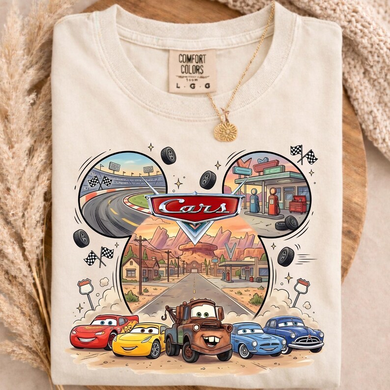 May include: Cream-colored t-shirt featuring a colorful graphic of the Disney Cars movie. The design includes race tracks, gas stations, and the Cars characters. The shirt has a Comfort Colors tag.