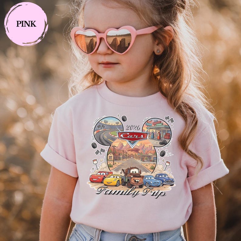 Puede incluir: Una camiseta rosa claro con un dise&ntilde;o de Cars, con el texto "2026 Cars Family Trip". El dise&ntilde;o presenta coches de dibujos animados, pistas de carreras y edificios. La camiseta la lleva una persona joven con gafas de sol rosas en forma de coraz&oacute;n.