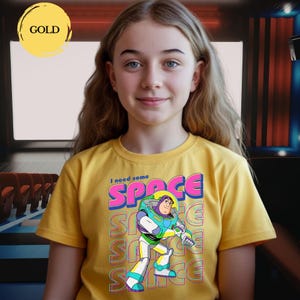May include: A young person wearing a yellow t-shirt with a graphic of Buzz Lightyear and the words "I need some SPACE". The design features a colorful illustration of the character with the word "SPACE" repeated in a retro font.