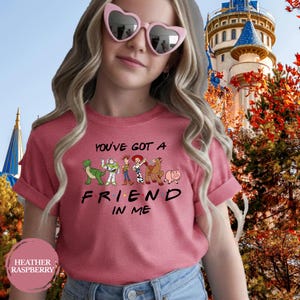 May include: Heather raspberry t-shirt with the text "YOU'VE GOT A FRIEND IN ME" and cartoon characters. The shirt features a design with a dinosaur, Buzz Lightyear, Woody, Jessie, Bullseye, and Hamm. The shirt is worn by a person wearing heart-shaped sunglasses.