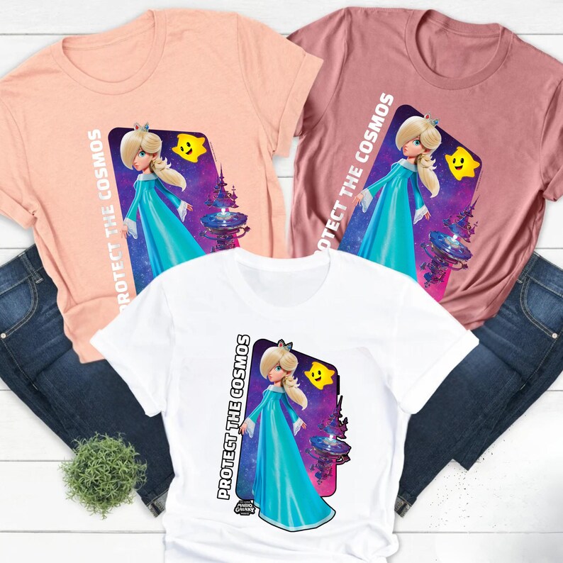 May include: Three short-sleeved t-shirts in peach, mauve, and white, each featuring a graphic of a character in a blue dress with the text "PROTECT THE COSMOS". The character is set against a purple and blue space-themed background.