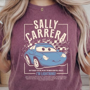 May include: A maroon t-shirt featuring a blue cartoon car with the text "SALLY CARRERA" above it. Below the car is the text "I'M LIGHTNING!" and other text. The design has a distressed, vintage look.