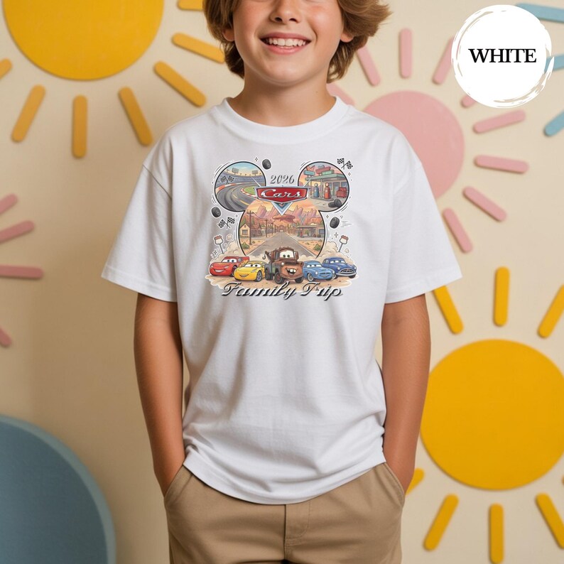 Puede incluir: Camiseta blanca con un gr&aacute;fico colorido de coches de dibujos animados y el texto "2026 Cars Family Trip". El dise&ntilde;o incluye im&aacute;genes de una carretera, una gasolinera y un paisaje des&eacute;rtico. La camiseta es de material suave.