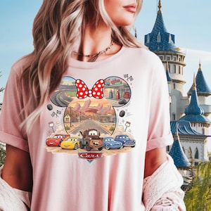 Pode incluir: Uma t-shirt rosa claro com uma impress&atilde;o gr&aacute;fica de personagens do filme Carros dispostos numa silhueta de orelhas do Mickey Mouse. O design inclui um la&ccedil;o vermelho com bolinhas brancas, uma cena de estrada no deserto e o log&oacute;tipo Cars.