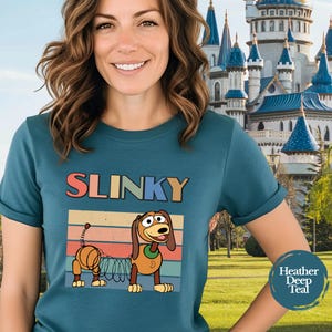 May include: A heather deep teal t-shirt featuring a graphic of a brown toy dog with a spring-like body. Above the dog, the word SLINKY is printed in colorful block letters. The design includes a retro-style sunset background with horizontal stripes.