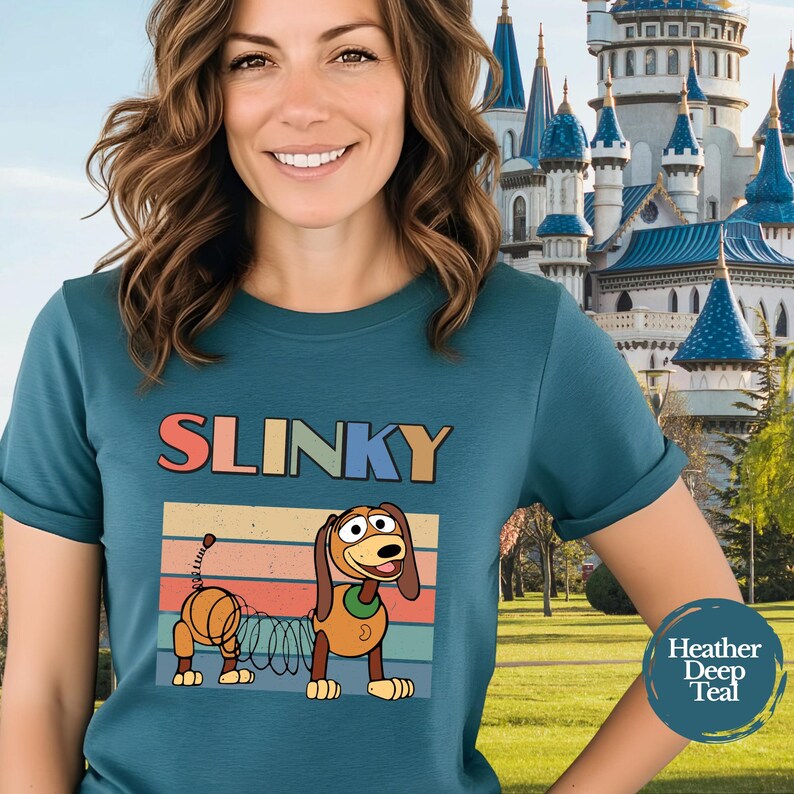 May include: A heather deep teal t-shirt featuring a graphic of a brown toy dog with a spring-like body. Above the dog, the word SLINKY is printed in colorful block letters. The design includes a retro-style sunset background with horizontal stripes.