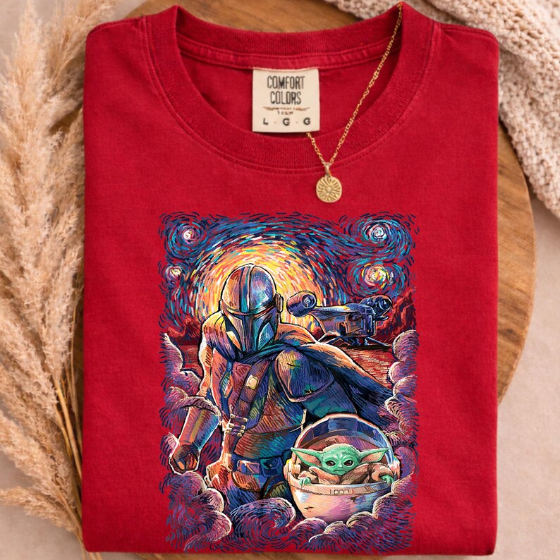 May include: Red Comfort Colors t-shirt with a graphic of The Mandalorian and Grogu in a starry night design. The shirt has a crew neck and short sleeves. A gold necklace with a sun pendant is draped over the shirt.