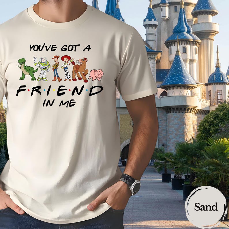 May include: Sand-colored t-shirt featuring the phrase "YOU'VE GOT A FRIEND IN ME" with cartoon character graphics. The design includes a dinosaur, Buzz Lightyear, Woody, Jessie, Bullseye, and Hamm. The shirt is a crew neck style.