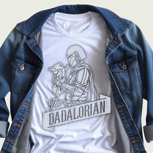 May include: White t-shirt with a grey graphic of a Mandalorian figure holding a small creature, and the text "The DADALORIAN" on a banner. The shirt is displayed on a blue denim jacket.