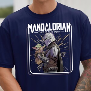 May include: Navy blue t-shirt featuring a graphic of The Mandalorian and Grogu. The design includes the text "MANDALORIAN - THIS IS THE WAY" in white. The illustration shows the Mandalorian holding Grogu, with a starburst background.