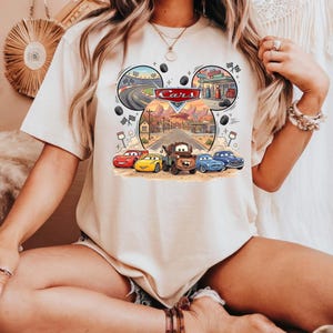 May include: Beige t-shirt featuring a colorful graphic of the movie "Cars." The design includes the word "Cars" in a red and white logo, along with illustrations of race cars, a gas station, and a desert landscape.