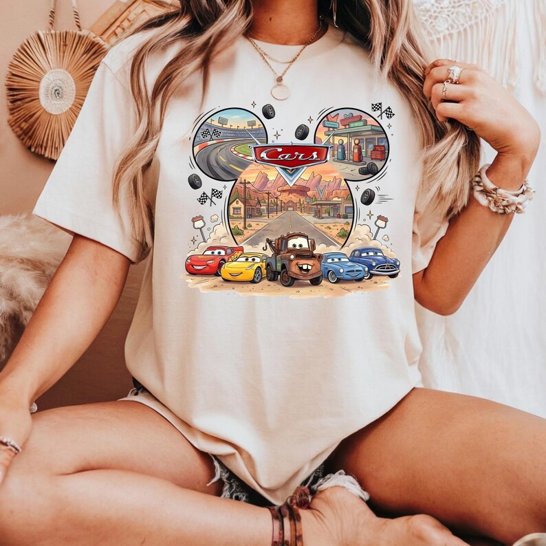 May include: Beige t-shirt featuring a colorful graphic of the movie "Cars." The design includes the word "Cars" in a red and white logo, along with illustrations of race cars, a gas station, and a desert landscape.