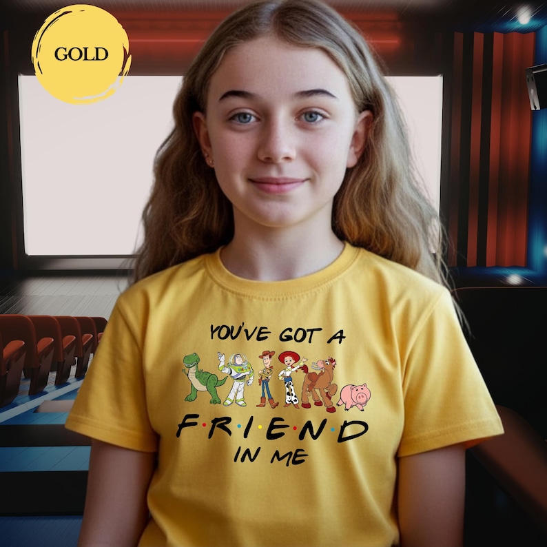 May include: A yellow t-shirt featuring the phrase "YOU'VE GOT A FRIEND IN ME" with cartoon characters. The shirt is worn by a person with long blonde hair. A gold circle with the word "GOLD" is in the upper left corner.