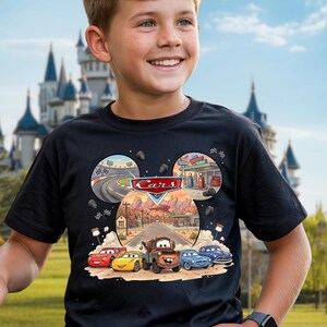 May include: Black t-shirt featuring a colorful graphic of characters from the movie "Cars." The design includes the word "Cars" in a red and white logo, and images of race cars and a desert landscape. The shirt is worn by a person.