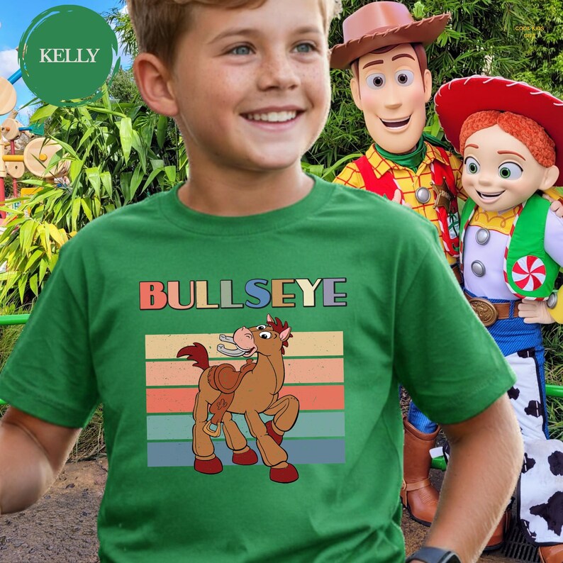 May include: A green t-shirt featuring a graphic of the horse character Bullseye from Toy Story. The design includes the text BULLSEYE in block letters above the horse, set against a background of horizontal stripes in muted retro colors.