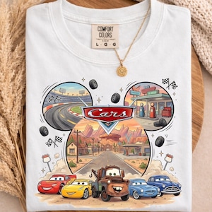 May include: White t-shirt featuring a colorful graphic from the animated movie "Cars." The design includes the "Cars" logo, race tracks, and various car characters. The shirt has a crew neck and short sleeves.