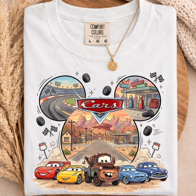 May include: White t-shirt featuring a colorful graphic from the animated movie "Cars." The design includes the "Cars" logo, race tracks, and various car characters. The shirt has a crew neck and short sleeves.