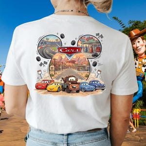 Pu&ograve; includere: Una t-shirt bianca con una stampa grafica dei personaggi del film Cars della Disney Pixar, tra cui Saetta McQueen, Cricchetto e altri. Il design include una scena stradale centrale, un circuito, una stazione di servizio e il logo Cars in rosso.