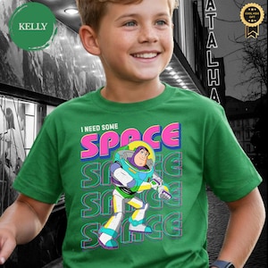 May include: Green t-shirt featuring a colorful graphic of Buzz Lightyear with the text "I NEED SOME SPACE". The design includes the word "SPACE" in vibrant pink, blue, and yellow, with repeated "SPACE" text in the background.
