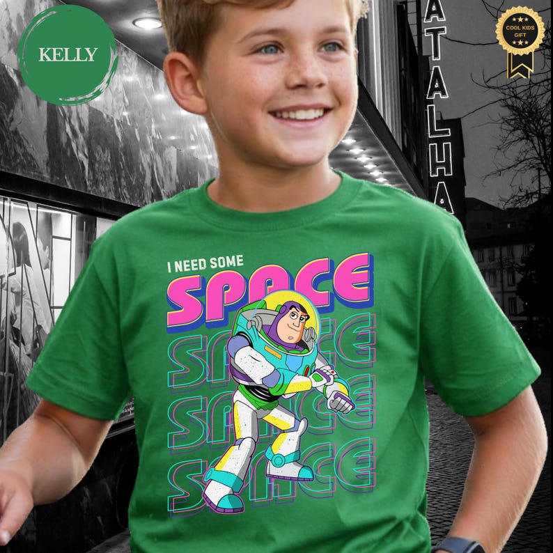 May include: Green t-shirt featuring a colorful graphic of Buzz Lightyear with the text "I NEED SOME SPACE". The design includes the word "SPACE" in vibrant pink, blue, and yellow, with repeated "SPACE" text in the background.