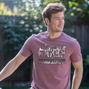 May include: Maroon crew neck t-shirt featuring a graphic design of Mandalorian helmets and the text "THE MANDALORIAN" in a silver metallic font. The design is centered on the front of the shirt.