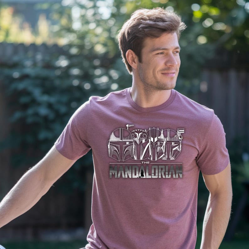 May include: Maroon crew neck t-shirt featuring a graphic design of Mandalorian helmets and the text "THE MANDALORIAN" in a silver metallic font. The design is centered on the front of the shirt.