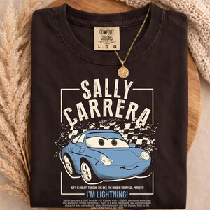 May include: Dark gray t-shirt featuring a blue cartoon car with the text "SALLY CARRERA" and "I'M LIGHTNING!". The shirt also has the text "COMFORT COLORS" and "L G G". A gold necklace with a sun pendant is also visible.