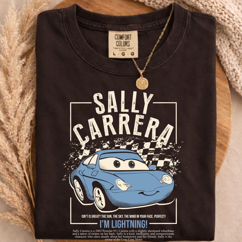 May include: Dark gray t-shirt featuring a blue cartoon car with the text "SALLY CARRERA" and "I'M LIGHTNING!". The shirt also has the text "COMFORT COLORS" and "L G G". A gold necklace with a sun pendant is also visible.