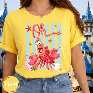 Pu&ograve; includere: Una t-shirt gialla a maniche corte con una stampa grafica di un granchio rosso e la scritta Oh in corsivo rosso. Il design include strisce verticali azzurre, varie stelle marine arancioni e conchiglie bianche sparse sul disegno.