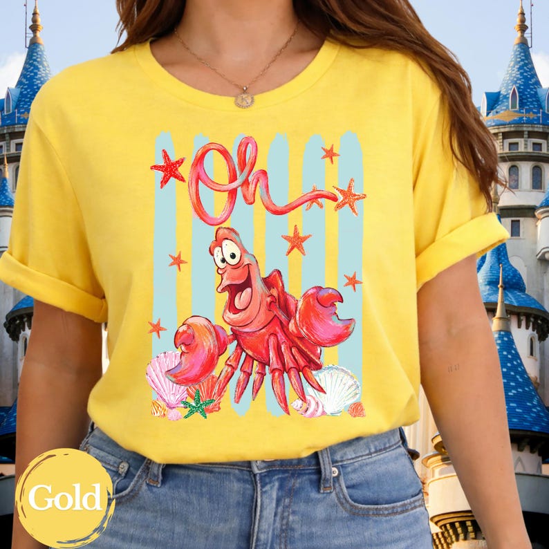 Pu&ograve; includere: Una t-shirt gialla a maniche corte con una stampa grafica di un granchio rosso e la scritta Oh in corsivo rosso. Il design include strisce verticali azzurre, varie stelle marine arancioni e conchiglie bianche sparse sul disegno.
