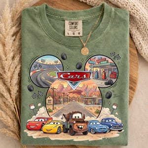 May include: Green t-shirt featuring a graphic of the Disney Cars movie. The design includes the Cars logo, race track, gas station, and the main characters: Lightning McQueen, Mater, and others. The shirt is a Comfort Colors brand.