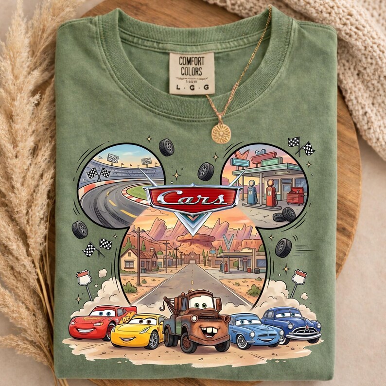 May include: Green t-shirt featuring a graphic of the Disney Cars movie. The design includes the Cars logo, race track, gas station, and the main characters: Lightning McQueen, Mater, and others. The shirt is a Comfort Colors brand.