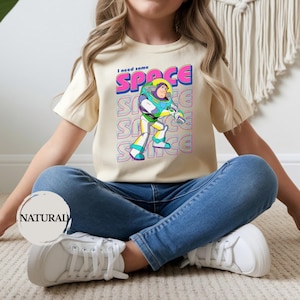 May include: A cream-colored t-shirt featuring a colorful graphic of Buzz Lightyear from Toy Story. The text "I need some SPACE" is above the character. The shirt is paired with blue jeans and white sneakers.