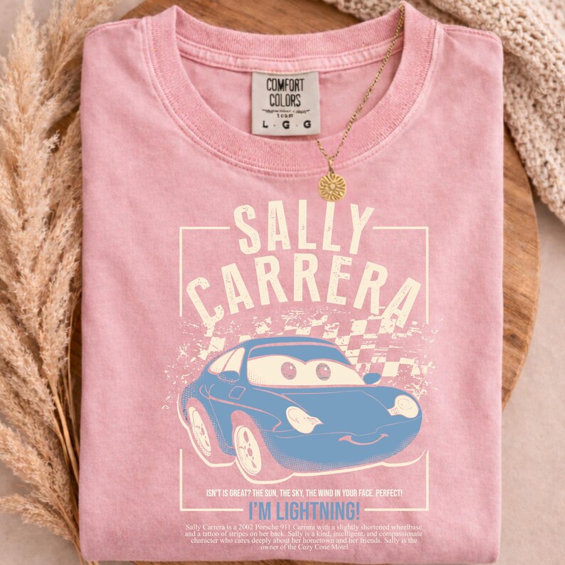 May include: A light pink t-shirt featuring a blue cartoon car with the text "SALLY CARRERA" and "I'M LIGHTNING!". The shirt has a crew neck and short sleeves. A gold necklace with a sun pendant is on the shirt.