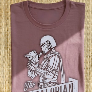May include: Mauve t-shirt with a white graphic of a Mandalorian character holding a small creature. The graphic includes the text "The Mandalorian" in a banner. The shirt is neatly folded.