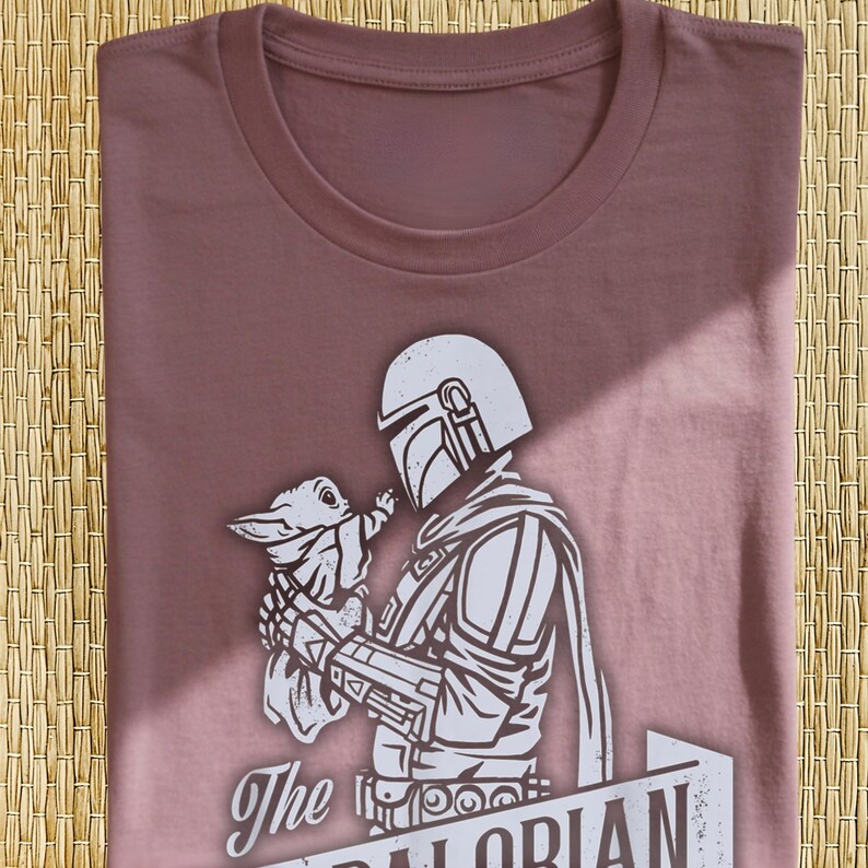 May include: Mauve t-shirt with a white graphic of a Mandalorian character holding a small creature. The graphic includes the text "The Mandalorian" in a banner. The shirt is neatly folded.