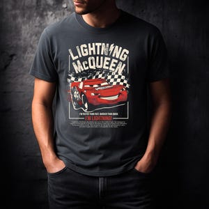 May include: Dark gray t-shirt featuring a graphic of Lightning McQueen from the Cars movie. The design includes the text "LIGHTNING McQUEEN" above a red race car with the number 95. Below the car is the phrase "I'M LIGHTNING!"
