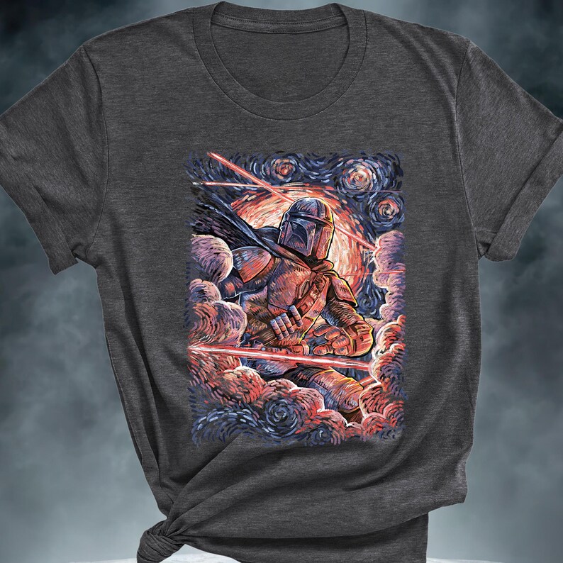 May include: Dark gray t-shirt featuring a stylized illustration of a Mandalorian warrior against a starry night sky. The artwork includes red laser beams and a vibrant color palette, creating a dynamic and artistic design.