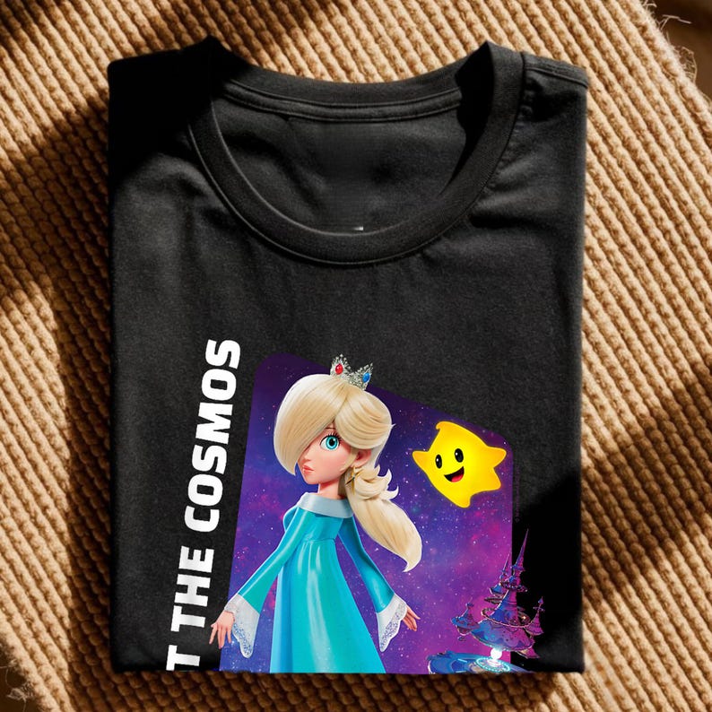 May include: Black t-shirt featuring a graphic of a character with blonde hair, a crown, and a blue dress, with a yellow star and the text "T THE COSMOS". The design is set against a purple and blue galaxy background.
