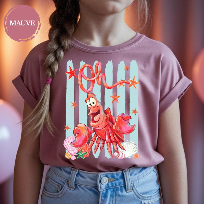 Pu&ograve; includere: Una t-shirt color malva a maniche corte con una stampa grafica di un granchio rosso dagli occhi grandi, circondato da pennellate verticali azzurre, stelle marine arancioni e conchiglie. La parola "Oh" &egrave; scritta in corsivo rosso sopra il granchio.