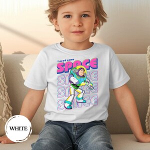 May include: White t-shirt featuring a colorful graphic of Buzz Lightyear with the text "I need some SPACE". The design includes the word "SPACE" in pink and blue, repeated vertically. The shirt is worn by a child.