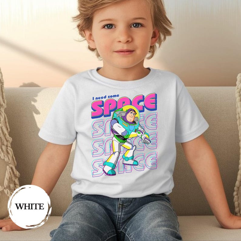 May include: White t-shirt featuring a colorful graphic of Buzz Lightyear with the text "I need some SPACE". The design includes the word "SPACE" in pink and blue, repeated vertically. The shirt is worn by a child.