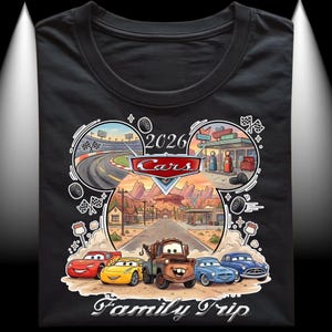 Puede incluir: Camiseta negra con un gr&aacute;fico colorido de la pel&iacute;cula Cars. El dise&ntilde;o incluye una pista de carreras, una gasolinera y el texto "2026 Cars Family Trip". La camiseta muestra a Rayo McQueen, Mate y otros personajes.