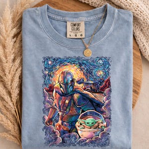 May include: Pale blue Comfort Colors t-shirt with a vibrant Starry Night-style illustration of The Mandalorian and Grogu. The shirt displays the Comfort Colors logo and size details. A gold necklace with a sun pendant rests on the shirt.