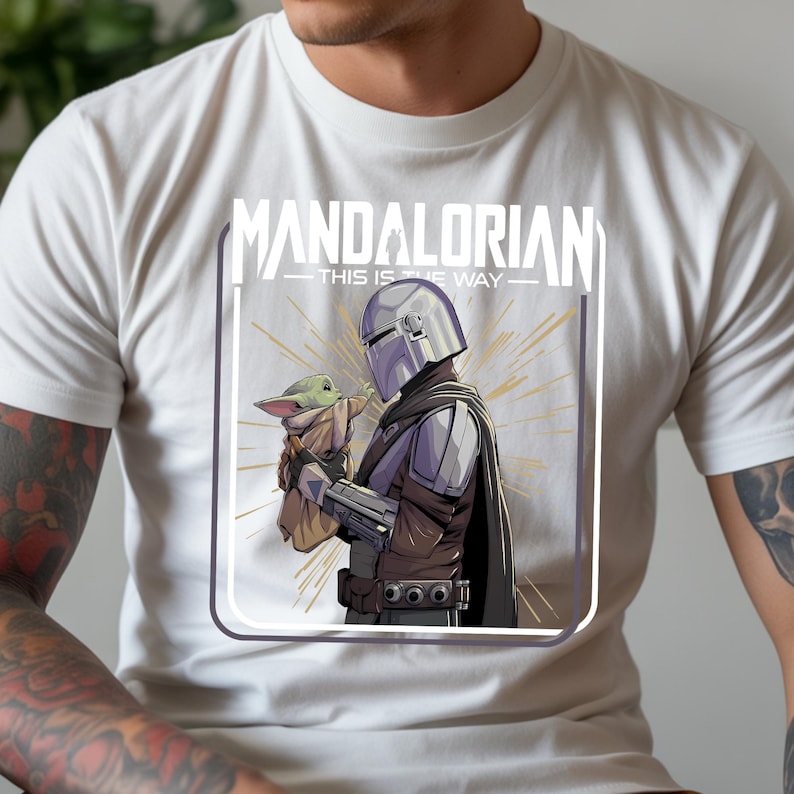 May include: White t-shirt featuring a graphic of The Mandalorian holding Grogu, with the text "MANDALORIAN - THIS IS THE WAY" in white. The design includes a silver helmet, brown and gray clothing, and a gold burst background.