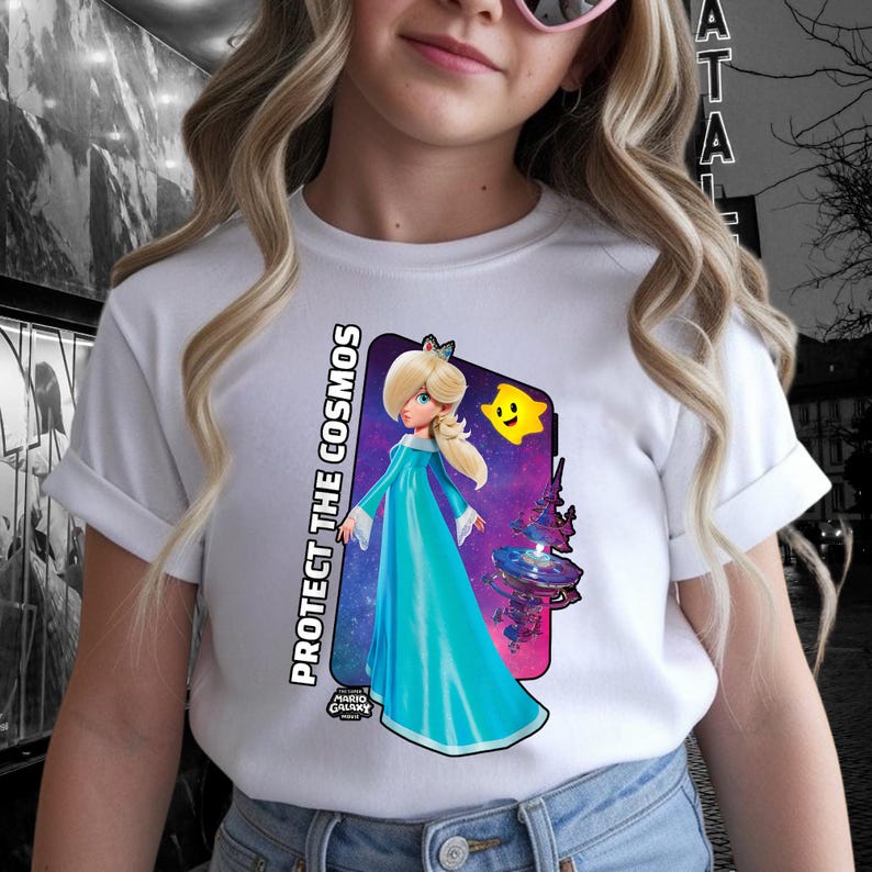 May include: White t-shirt featuring a graphic of a character in a blue gown with the text "PROTECT THE COSMOS". The graphic includes a yellow star and a space-themed background. The shirt is worn by a person.